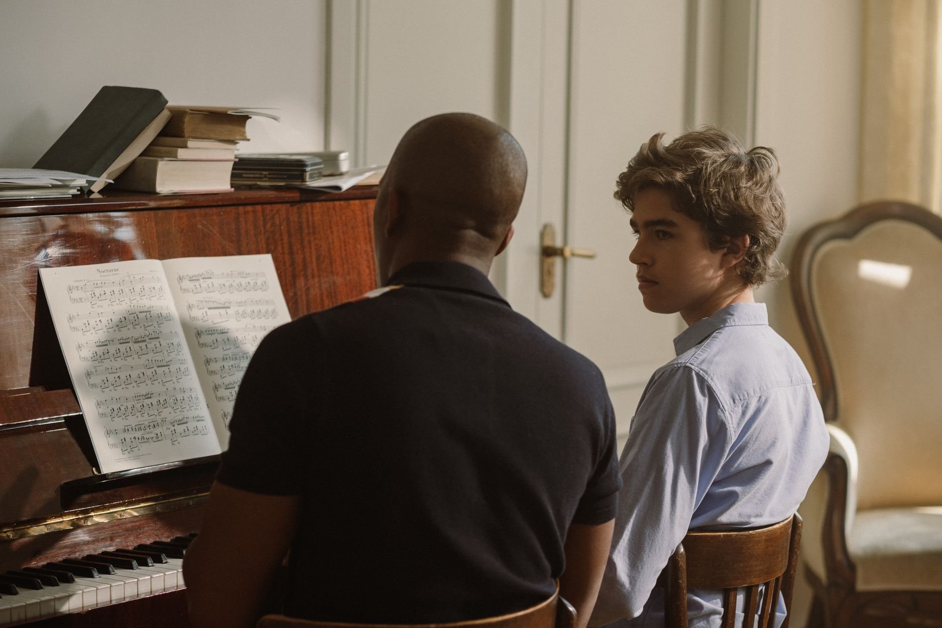 Un profesor de música guía a un alumno durante una clase de piano en una sala con luz cálida.
